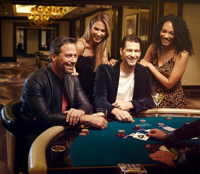 Two men sitting at a poker table with two women behind them